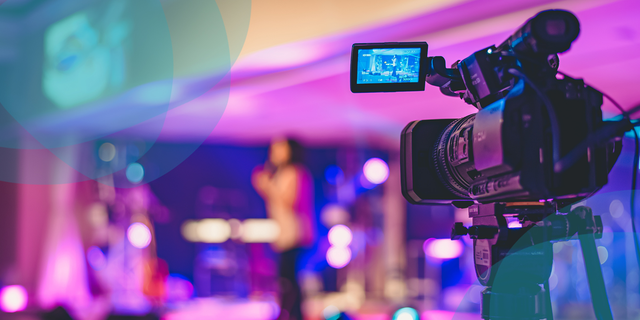 A video camera is set up to show a woman at a podium on stage. This shows the set up of an in-person worship service.