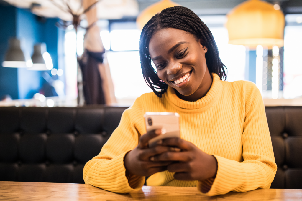 Young African American woman in a yellow turtleneck sits at a table, smiling and holding her smart phone after downloading the Givelify app for mobile and online giving.